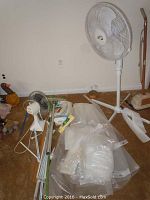 Photo showing white pedestal fan, smaller white desk fan, clear large plastic wrap rolls, assorted mops, some filters and cleaning accessories on carpeted floor.