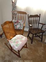 Set of three chairs: two wooden rocking chairs and one wooden armchair. One rocking chair has a cane backrest and patchwork cushion, another has vertical wooden slats and floral cushion, third is a dark wood chair with armrests.