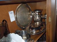Photo showing brass silver plated pitcher, large tray, two goblets, serving platter and utensils inside cabinet.