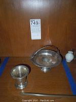 Photo showing two silver items: a footed bowl with detailed rim and a smaller matching vase or cup on a wooden surface with a label behind.