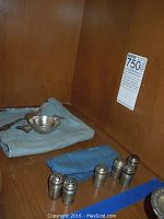 Six small sterling silver shakers arranged on a wooden surface, some placed near blue cloth pieces, tagged as 750 grams total silver weight.