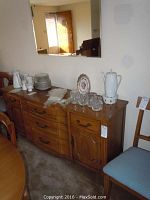 Medium dark wooden buffet with various dishware on top, placed below the mounted mirror. Buffet shown with three drawers at center and cabinet doors on both sides.