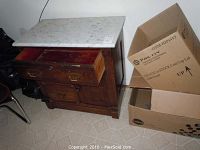 Front and side view of the vintage wooden wash stand showing marble top and two drawers partially open with general wear and boxes beside it.