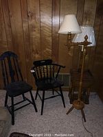Two black wooden chairs, standing adjustable lamp with two shades, and wooden lamp table against wood panel wall, on carpeted floor.