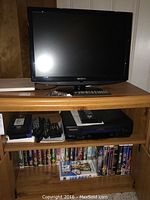 Insignia 21-inch TV on top of wooden cabinet with Panasonic VCR, VHS tapes, and remotes arranged on lower shelves