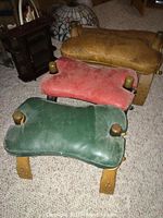 Three leather and wood stools placed on carpet floor showing colors tan, green, and red with worn leather and brass damage