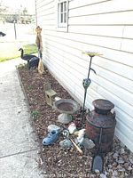 Outdoor decor items arranged along a building's siding including tall black goose decoys, stone bird bath, rusty metal milk jug, garden gnomes, small gardening tools, and flower garden stakes.
