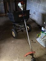 Photo showing Agri Fab towable seeder with black cracked plastic container and rust on frame, alongside Stihl FS55 weed wacker with white motor and orange safety guard