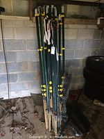 Photo showing 11 green metal fence posts bundled together upright against a cinder block wall in a garage or shed. Yellow clips attached on posts visible. Dust and dirt on the ground.