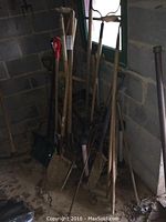 Full view of multiple used gardening tools standing in a corner of a concrete block wall basement or storage area, showing shovels, rakes, pick axes, and other handles leaning against the wall.
