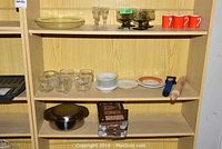 Three shelf section with glassware including drinking glasses with globe motif, crystal vase, stacked small plates, orange cups, and a wooden rolling pin.