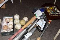 Photo showing assortment of baseball bats, baseballs in box and loose, golf balls in package, and a dart set with case.