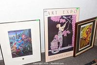 Four framed art prints leaning against a wall, showing Tom Palmer superhero art, Paula Mae Green art expo poster, a fantasy art print, and a Route 66 decor piece.