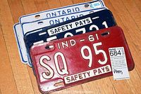 Six license plates in a pile showing two blue and white Ontario handicap plates, two Indiana 'Safety Pays' plates from 1960 and 1961, and two white Ontario plates dated December 8.