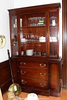 Front view of wooden china cabinet with glass display shelves showing glassware and dishware inside; large drawers and two side cabinet doors below; shows wear and detailing on top crown molding.