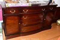Front view of wooden dining room server showing bow front curvature, drawers, cupboard doors, and brass-tone handles.