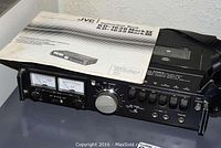 Front view of black JVC portable cassette deck recorder featuring metal knobs, dual VU meters, and control buttons with instruction manual placed on top.
