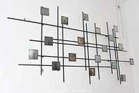 Full view of the large metal wall art mounted on the wall, showing intersecting black rods with attached square metal pieces.