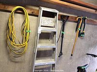 Photo showing the yellow electrical extension cord, aluminum ladder, and garden shovel and pickaxe hanging on the wall