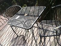 Photo showing black wrought iron square patio table with mesh top and two matching chairs on wooden deck, highlighting overall set condition and design.