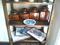 Four graduated brown glazed ceramic kitchen canisters labeled Flour, Coffee, Sugar, and Tea with black tags, placed on a wood shelf alongside a Bermuda decorative wall plate and four wood-framed recipe prints.