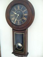Full view of Bulova wooden wall clock showing round dial with Roman numerals, clock hands, and pendulum housing below.