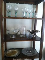 Shelf with multiple glassware and silver plate items including eight wine glasses with teal stems, two clear drinking glasses, a clear glass pitcher, footed silver plate and copper fruit bowl, clear glass bowl with silver plate foot, a small clear glass bowl, and a heavily tarnished silver plate tray and clear glass plate on bottom shelf.