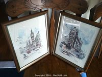 Pair of framed Jan Korthals prints showing Big Ben and Tower Bridge, positioned upright on wooden chairs.