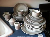 Photo showing a full set of stacked dishes including dinner plates, cups, bowls, and serving tray arranged on a blue surface.