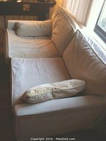 Front-side angled view of the sand colored velour loveseat with plush cushions and bolster pillow, showing fabric texture and wear.