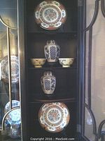 View of all items in a cabinet showing the two ginger jars with lids on wooden bases, two floral motif plates, and two rice bowls arranged in three shelves.