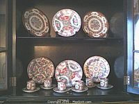 Shelf display showing six hand-painted Chinese porcelain plates on top shelf and six matching tea cups with saucers on bottom shelf, arranged in pairs. Plates and cups have colorful traditional designs with floral and figure motifs, gold lines on plates visible.