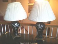 Pair of black metal table lamps with mother of pearl inlays on bronze-tone bases, each topped with white pleated silk lampshades.