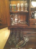Full view of the lot showing clear glass pitcher, cordial and aperitif glasses, small creamer and two leaf-shaped glass dishes arranged on wooden furniture.