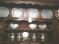 Eight pewter plates displayed on upper and middle shelves with four pewter mugs on bottom shelf inside wood cabinet.