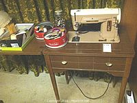 Singer electric sewing machine in wooden cabinet with extendable table and various sewing accessories arranged on the side including buttons and thread.