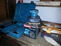 Blue Makita finishing sander sitting on box on a wooden shelf with carrying case behind