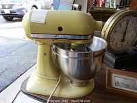 Vintage yellow KitchenAid mixer with stainless steel bowl attached, power cord visible, outside setting near window.