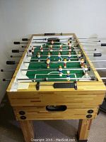 Full view of wooden foosball table showing playing surface with 8 metal rods and plastic foosball men in red and blue colors, green field, and wood laminated sides and legs.
