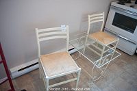 White wrought iron side table with clear glass top between two white wrought iron chairs with wood and wicker seats, placed on tiled floor next to stove.