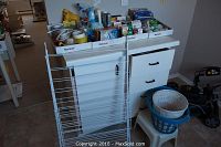 Overview photo showing a white foldable drying rack, white plastic step stool holding a blue laundry basket, various household items on white cabinet with drawers and boxes of supplies on top.