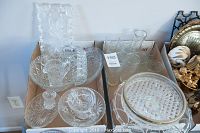 Two trays of clear glass items including serving plates, trays, cake plate, and drinking glasses arranged together.