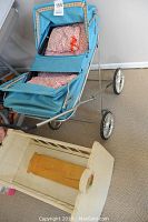 Photo of vintage blue doll carriage with metal frame and four spoked wheels, showing side profile and part of the rocking cradle next to it