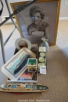 Photo showing framed portrait of Princess Diana with several decorative tins, ship models and a ceramic figurine arranged on a carpeted floor, under glass.