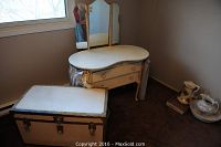 Photo of antique white two drawer vanity with tri-fold mirror and skirt along with a vintage white steamer trunk placed beside it on the floor near a window.