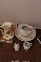 Two ceramic pitchers and wash basin set on table, showing the overall lot including large cream floral pitcher and white with red floral basin and ceramic pieces.