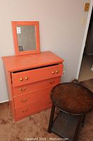 Full view of orange painted dresser with four drawers and a separate matching mirror, next to a dark oak round side table.