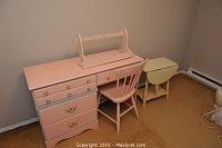 Pink painted desk with several drawers, matching pink chair, small cream colored flip-top side table, and cream wall shelf with hooks in a room with brown carpet
