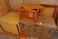 Photo displaying a silver metal steamer trunk, a large wooden box, and a smaller wooden box with lid placed in a room corner against wood paneling and a concrete wall.