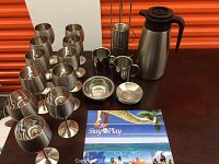 Overview of set on dark surface with orange background showing eight stainless steel wine goblets, three coffee cups, small bowl, saucer, a stainless steel utensil holder with four stirrers, and a stainless steel carafe with black plastic lid and handle.